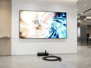 Groot digitaal beeldscherm aan witte wand in moderne lobby, met mediaspeler en HDMI-kabel op de vloer eronder.