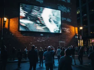 Groot digitaal reclamescherm op bakstenen muur met storingen, omstanders kijken omhoog op drukke stadsstraat, elektrisch blauwe en amber tinten.