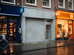 Grijze, naamloze winkel tussen levendige, kleurrijke gevels op een Nederlandse winkelstraat met kobaltblauwe en oranje uithangborden.